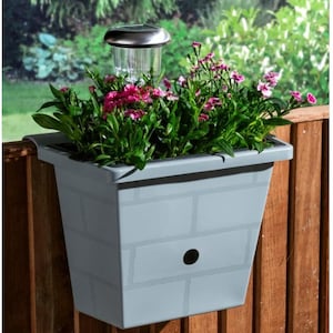 Könnte beinhalten: Grauer Pflanzkasten mit Ziegelmuster, gefüllt mit rosa Blüten und grünem Laub, an einem Holzzaun befestigt. Eine solarbetriebene Leuchte sitzt oben auf dem Pflanzkasten und verleiht dem Außenbereich eine dekorative Note.