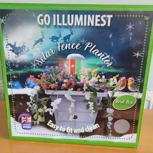 以下が含まれることがあります： Go Illuminestソーラーフェンスプランターの長方形の製品ボックス。箱には、色とりどりの花、ソーラーライト、2羽のコマドリで満たされた白いプランターのグラフィックが描かれています。テキスト：Go Illuminest、Solar Fence Planter、Bird Box。