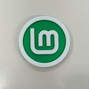 Pode incluir: Um emblema circular com borda branca e centro verde, apresentando um logótipo branco estilizado com as letras minúsculas L e M. O design tem uma superfície texturizada, típica de objetos impressos em 3D, exibindo o logótipo Linux Mint.
