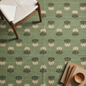 May include: A sage green area rug featuring a repeating pattern of stylized cream and dark green flowers. The rug has a fringed edge and is displayed with a wooden chair with a woven seat and a stack of books topped with a small wooden bowl.