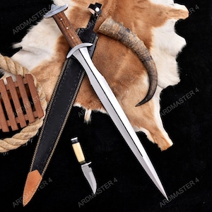 Puede incluir: Colección de armas blancas, que incluye una espada grande con empuñadura de madera y un cuchillo más pequeño. La espada está enfundada en una vaina de cuero negro. También hay un cuerno curvado y un elemento decorativo de madera.