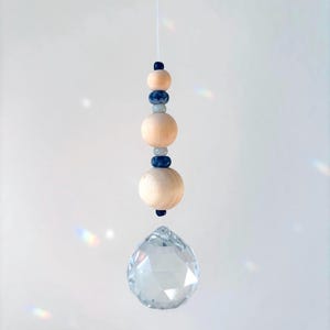 Könnte beinhalten: Ein hängender Kristall-Sonnenfänger. Am unteren Ende hängt eine klare, facettierte Glaskugel, darüber Holz- und blaue Perlen, auf einer weißen Schnur aufgereiht. Er soll Sonnenlicht einfangen und Regenbogen erzeugen.