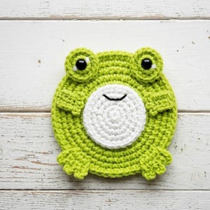 May include: A handmade, lime green crochet frog coaster with a white circular face and black eyes. The coaster has a round shape with small arms and feet. The frog coaster is designed to protect surfaces from drink condensation.