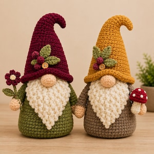 May include: Two handmade crochet gnomes featuring textured beards and tall pointed hats. One gnome wears a deep burgundy hat with a flower, while the other wears a mustard yellow hat with a red mushroom. Both have green and brown bodies.