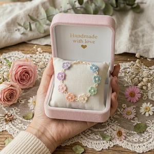 May include: A delicate crochet flower bracelet featuring pastel-colored petals and small white beads, displayed on a cream-colored cushion inside a pink velvet jewelry box with the text Handmade with love and a heart icon printed inside.