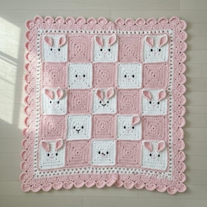 May include: A handmade crochet blanket featuring a repeating pattern of bunny faces in white squares against a pink background. The blanket has a scalloped pink border and is made of soft yarn. The overall design is a charming, whimsical style.