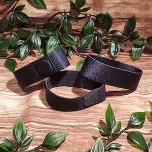 Könnte beinhalten: Drei marineblaue elastische Bänder mit Klettverschlüssen. Die Bänder sind auf einer Holzoberfläche mit grünen Blattranken im Hintergrund angeordnet. Die Bänder scheinen aus einem gewebten Stoff zu bestehen.