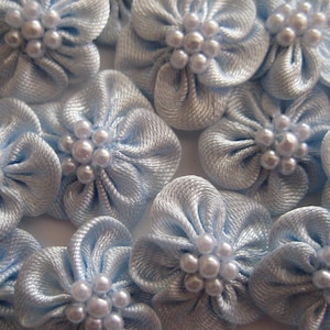 May include: A collection of light blue satin flowers with white pearl centers. The flowers are arranged in a close-up view, showcasing their delicate details.