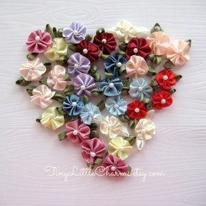 May include: A collection of handmade fabric flowers in various colors, including pink, red, blue, purple, and cream. The flowers are arranged in a heart shape on a white wooden surface. Tiny Little Charms.etsy.com