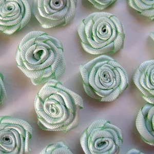 May include: A collection of 15 light green satin ribbon rosettes. The rosettes are approximately 1 inch in diameter and have a delicate, ruffled appearance.