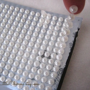May include: A sheet of white pearl-like stickers on a black surface. The stickers are arranged in a grid pattern. A finger is pressing down on one of the stickers.