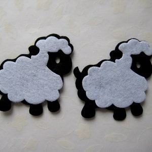 May include: Two black felt sheep with white felt wool. The sheep are standing on a white surface.
