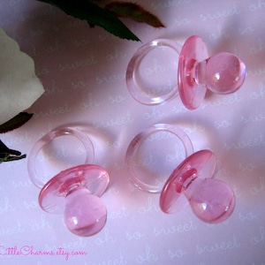 May include: Four pink plastic baby pacifier favors for a baby shower or party. The pacifiers are clear with a pink tint and have a ring around the center.
