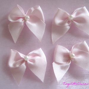 May include: Four light pink satin bows, each with a small, white pearl in the center. The bows are arranged on a light pink background. The bows are likely for crafts or embellishments.