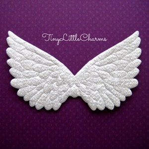 May include: White glitter angel wings with a feathery texture. The wings are on a purple background with a white text overlay that reads "TinyLittleCharms".