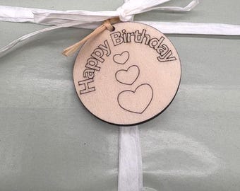 Może przedstawiać: Okrągła drewniana zawieszka na prezent z napisem "Happy Birthday" i trzema sercami. Zawieszka jest przymocowana do zapakowanego prezentu białą wstążką. Prezent jest owinięty w jasną zieloną papier.