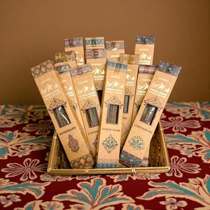 May include: A collection of Himalayan Masala incense sticks in decorative cardboard packaging. The boxes, in shades of brown and beige, feature mountain and floral designs, with labels indicating scents like Rosemary, Lavender, and White Sage. The incense is displayed in a woven basket.