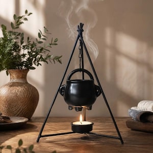 May include: A black ceramic cauldron oil burner suspended from a triangular metal tripod stand. A small tea light candle sits in a holder at the base, heating the cauldron and releasing a thin wisp of white smoke into the air above the vessel.