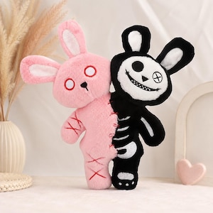 May include: A plush toy featuring two conjoined rabbit halves. The left side is soft pink with red stitched X details and circular eyes. The right side is black with a white face, a wide stitched grin, a circular eye, and a white ribcage design.