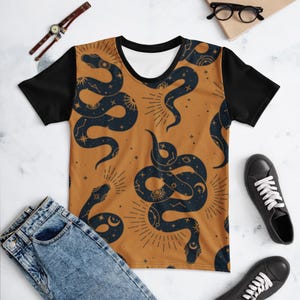 May include: A short-sleeve t-shirt with a black crew neck and black sleeves. The main body features a repeating pattern of dark navy blue snakes on a burnt orange background, accented with white celestial symbols like stars, suns, and moons.
