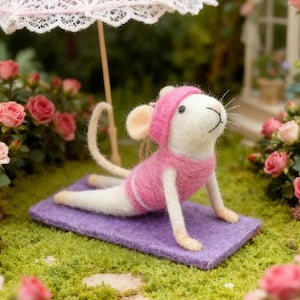 May include: A felt mouse in a pink outfit and hat doing yoga on a purple mat under a white lace parasol. The mouse is surrounded by pink roses and green grass, creating a whimsical scene.