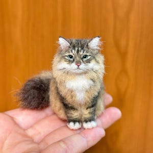 Peut inclure: Figurine miniature de chat détaillée avec fourrure marron, beige et blanche, yeux verts et une queue touffue. Le chat est assis sur une main, mettant en valeur le savoir-faire de l'article fait à la main.