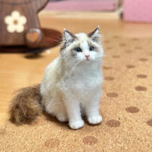 Op de afbeelding: Handgemaakte, pluizige wit-bruine Ragdoll kattenfiguur met blauwe ogen. De kat zit rechtop, met zijn staart opzij gekruld. Het figuur is gemaakt van zacht, getextureerd materiaal en is gedetailleerd afgewerkt.