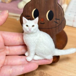 Op de afbeelding: Handgemaakte witte kattenfiguur met gele ogen, zittend op een hand. De kat is gemaakt van een zacht, getextureerd materiaal. Achter de kat staat een houten katvormige decoratie met een lachend gezicht.