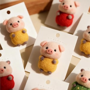 May include: Handmade felt pig brooches in various colours, including yellow and red. Each pig has a pink snout, ears, and a matching outfit. The brooches are attached to white cards, ideal for gifting or display.