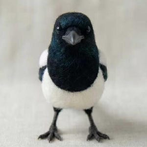 Puede incluir: Una figura de urraca hecha con fieltro de aguja que presenta cabeza, cuello y alas negras con plumaje blanco en el pecho y los costados. El ave tiene un pico oscuro y puntiagudo, ojos negros pequeños y patas oscuras, finas y articuladas.