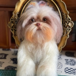 Puede incluir: Escultura artesanal de un perro con un pelaje blanco y canela esponjoso, ojos marrones y un hocico color melocotón. El perro está posicionado frente a un espejo con marco dorado, creando una exhibición encantadora. La escultura está hecha de materiales suaves y texturizados.