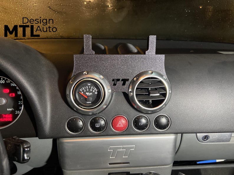 May include: Black dashboard with a custom gauge and vent setup. The gauge reads in bar and has a red needle. The vent is circular with silver trim. The text "TT" is visible. The text "Design MTL Auto" is in the upper left corner.