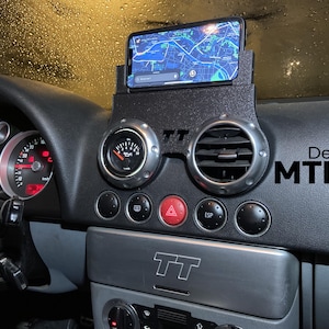 Peut inclure: Tableau de bord de voiture noir avec un support de téléphone affichant une carte. Le tableau de bord comporte des jauges et des bouches d'aération cerclées d'argent. Le texte "Design MTL Auto" est visible sur le côté droit.