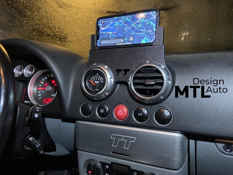 May include: Black car dashboard with a phone holder displaying a map. The dashboard has silver-rimmed gauges and air vents. The text "Design MTL Auto" is visible on the right side.
