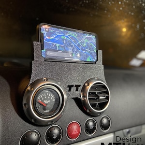 May include: Black car dashboard with a phone holder, gauge, and air vent. The phone displays a map. The dashboard has the text "TT" and "MTL Auto". The gauge reads "bar" and "oil".