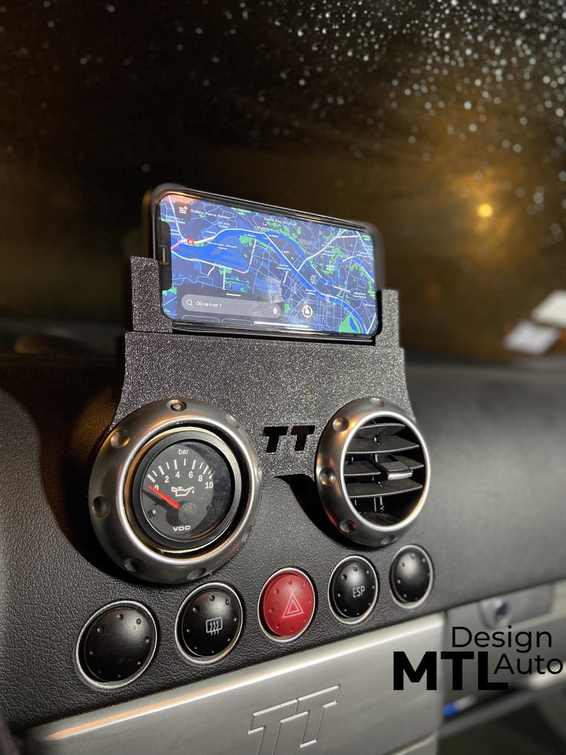 May include: Black car dashboard with a phone holder, gauge, and air vent. The phone displays a map. The dashboard has the text "TT" and "MTL Auto". The gauge reads "bar" and "oil".