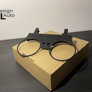 May include: A black plastic car part with two circular openings and the letters "TT" in the center. The part is resting on a cardboard box. The text "Design MTL Auto" is in the upper left corner.