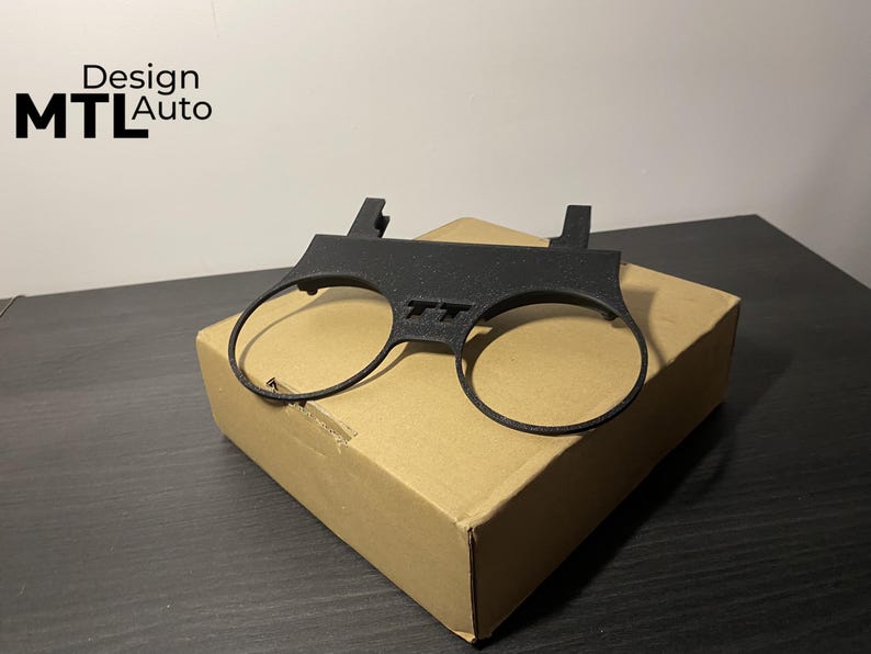 May include: A black plastic car part with two circular openings and the letters "TT" in the center. The part is resting on a cardboard box. The text "Design MTL Auto" is in the upper left corner.