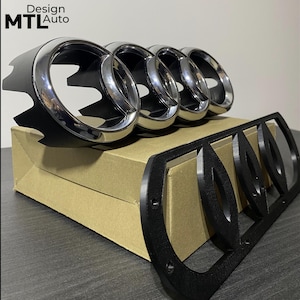Puede incluir: Un conjunto de cuatro anillos cromados, un emblema de coche y un marco negro. Los anillos están montados sobre una caja de cartón. El texto "Design MTL Auto" es visible en la esquina superior izquierda.