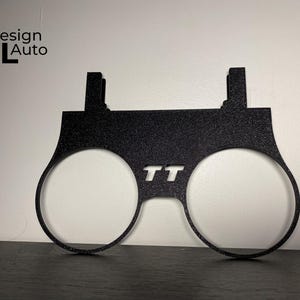 May include: Black metal automotive part with two circular openings and the letters "TT" cut out. The part has a textured surface and two rectangular tabs at the top. The text "Design MTL Auto" is in the upper left corner.