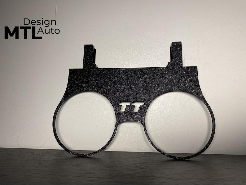 May include: Black metal automotive part with two circular openings and the letters "TT" cut out. The part has a textured surface and two rectangular tabs at the top. The text "Design MTL Auto" is in the upper left corner.