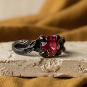 May include: A black ring with a large, round, red gemstone. The ring has a floral design on the band and around the gemstone. The ring is set on a light-colored, textured surface, with a mustard-colored fabric in the background. Jewelry.