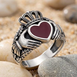May include: A silver Claddagh ring featuring two hands holding a central heart with a deep red inlay, topped with a crown. The metal band displays intricate leaf detailing on the sides, showcasing a polished finish with darkened recessed areas.