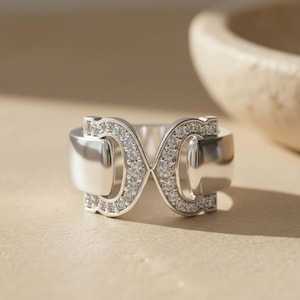 May include: A silver ring featuring a double infinity symbol, embellished with small gemstones. The ring is displayed on a neutral surface, with a blurred bowl visible in the background.