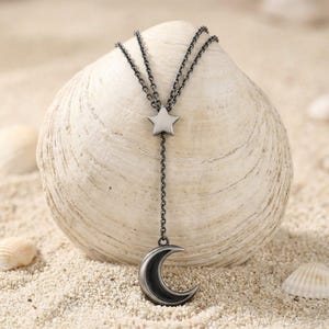 Può includere: Una collana lariat in metallo scuro caratterizzata da una doppia catena che si collega a un ciondolo a forma di stella a cinque punte, che si estende poi in una catena singola terminante con un pendente a mezzaluna, con finitura grigio scuro.