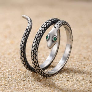 May include: A silver-toned wrap ring shaped like a coiled snake with a textured scale pattern covering the band. The snake head features two small, round, vibrant green eyes, and the tail curves upward at the end of the metal band design.
