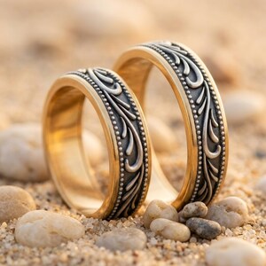 May include: Two gold wedding bands with intricate silver filigree designs. The bands are set against a sandy background with small pebbles, creating a warm, romantic scene. The rings feature a detailed pattern of swirling leaves and beaded edges.