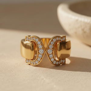 Könnte beinhalten: Ein goldener Ring mit einem doppelten Unendlichkeitszeichen, besetzt mit kleinen, funkelnden Diamanten. Der Ring hat eine polierte Oberfläche und ist vor einem neutralen Hintergrund platziert, mit einer unscharfen Schale im Hintergrund.