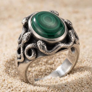 May include: A silver-toned ring featuring a central oval-shaped green malachite stone with concentric banding. The band and setting are sculpted with multiple detailed snake figures that wrap around the stone and along the sides of the ring.