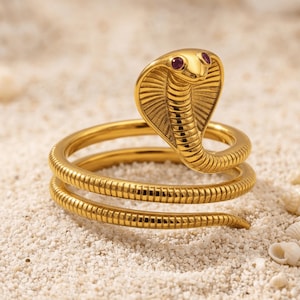 May include: A gold-toned metal ring shaped like a coiled cobra snake. The snake features a raised hood with detailed scale textures and two small, round, deep pink gemstone eyes. The body of the snake wraps around to form the band of the ring.